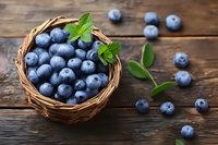 Fresh ripe blueberries in wicker basket with mint leaves on rustic wooden background, scattered berries and green foliage create natural composition.