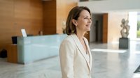 Professional businesswoman in beige suit smiling confidently in modern corporate lobby with reception desk and contemporary interior design.