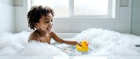 Happy African American toddler girl enjoying bubble bath with yellow rubber duck toy in modern white bathroom. Child bathing, hygiene, childhood joy.