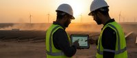 Two male engineers in safety gear discussing wind farm project on tablet at sunset in desert renewable energy site for construction planning.
