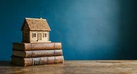 Wooden house model sitting on stack of old books against blue background representing real estate education and property investment knowledge.