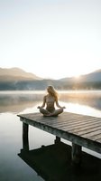Young Caucasian woman meditating in lotus pose on wooden dock at misty mountain lake during golden sunrise, embodying peace and mindfulness.