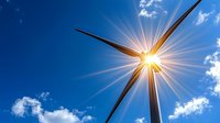 Wind turbine blade against bright blue sky with sunburst effect and white clouds, representing renewable energy and sustainable power generation solutions.