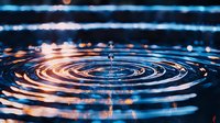 Water drop falling into dark liquid creating concentric ripple circles with orange and blue light reflections on surface in macro photography.