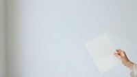 A person with light skin tone holds a blank white sheet of paper against a neutral wall, ideal for mockups, presentations, and advertising campaigns.
