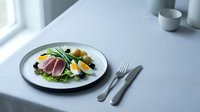 Nicoise salad with seared tuna, boiled eggs, green beans, olives and potatoes served on a white plate with cutlery on a light gray linen tablecloth.