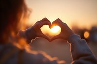 Woman making heart shape with hands at sunset capturing warm golden light through fingers creating romantic silhouette against blurred background.