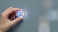 Hand holding a glowing social media camera icon button with neon blue light effect on gray background, digital technology concept.