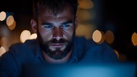 Bearded man illuminated by blue screen light in dark room with bokeh background, dramatic moody portrait showing serious concentrated expression.
