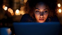 Young mixed-race woman with curly hair working late on laptop computer in dark office with warm bokeh lights creating atmospheric evening workspace ambiance.