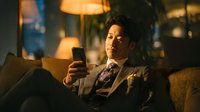 Professional Asian businessman in suit relaxing on sofa using smartphone in warm evening lighting for corporate lifestyle and technology concepts.