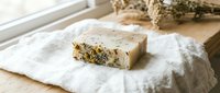 Handmade soap bar with dried herbs and botanicals on white linen cloth, natural skincare product for wellness and beauty routines.