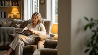 Middle-aged woman with blonde hair reading a book while relaxing in a comfortable armchair by a sunny window in a modern living room with warm lighting and home decor.