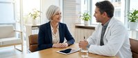Professional mature Caucasian businesswoman in navy suit consulting with middle-aged male doctor in white coat at modern office desk during healthcare meeting.