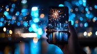 Smartphone capturing fireworks over city skyline at night, framed by blurred blue bokeh lights, reflecting on water surface. Urban photography through device screen.