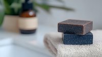 Handmade soap bars in brown and blue colors stacked on white cotton towel, natural skincare product photography with soft bokeh background and plant decor.