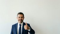 Middle-aged man with dark hair and beard, Mediterranean ethnicity, wearing navy business suit and tie, giving thumbs up gesture against white background. Isolated stock photo suitable for corporate, business success, and professional website design.