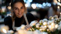 Young female florist in black apron arranging white flowers with soft bokeh lights creating magical atmosphere in flower shop. Professional at work with gentle smile.