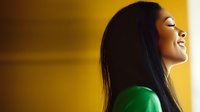 Young Asian woman with long dark hair laughing with eyes closed against yellow wall background, wearing green top, expressing joy and genuine happiness in profile view.