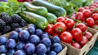 Fresh organic produce in wooden crates including ripe plums, blackberries, cherry tomatoes, and cucumbers arranged at farmers market or grocery store display.