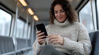 Young woman in cozy beige sweater smiling while using smartphone on public transport, looking at mobile screen with happy expression. Modern lifestyle.