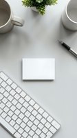 Minimalist white desk workspace with blank business card, keyboard, coffee cups, pen and green plant for branding mockup and corporate identity design.