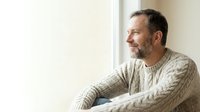 Middle-aged man with dark hair and beard wearing cable-knit sweater, gazing thoughtfully out window in natural daylight. Suitable for lifestyle, wellness, and contemplation themes.