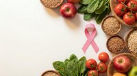 Pink ribbon surrounded by healthy foods including fresh vegetables, fruits and whole grains on white background promoting breast cancer awareness and nutrition.