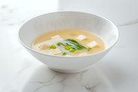 Traditional miso soup with soft tofu cubes and fresh green onions served in white ceramic bowl on marble surface, Japanese cuisine preparation.