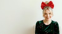 Young woman with blonde hair wearing a large red sequined bow headband and dark green velvet top, smiling at camera against neutral background. Suitable for holiday, fashion, and festive design projects.