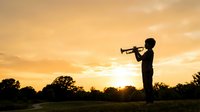 Silhouette of male musician playing trumpet outdoors at golden sunset in open field, suitable for music, inspiration, and performance concepts.