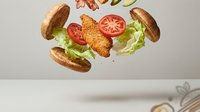 Crispy fried chicken sandwich with fresh lettuce and tomato slices suspended in mid-air, capturing dynamic food photography for culinary and restaurant marketing.