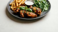 Glazed chicken wings with golden fries and fresh arugula salad served on dark plate with creamy dipping sauce for restaurant menu and food photography.