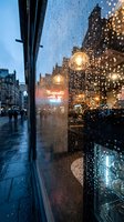 Rainy evening city street view through wet glass window with water droplets reflecting warm streetlights and urban architecture creating atmospheric mood.