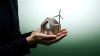 Businessperson holding miniature eco house model with solar panel and wind turbine representing sustainable energy and green building concepts.