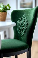 Green velvet dining chair with embroidered leaf pattern detail, photographed in soft natural light against white interior background, showcasing elegant home decor.