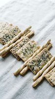 Assorted seeded crackers and breadsticks arranged on white linen fabric, featuring poppy seeds, sesame seeds, and rosemary herbs for food styling and culinary content.