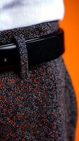 Close-up view of black leather belt with textured tweed fabric in black and orange speckled pattern against vibrant orange background, showing fashion detail.
