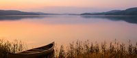 Wooden boat on tranquil lake shore at sunset with golden reflections and mountain silhouettes creating peaceful waterscape for relaxation and nature themes.