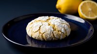 Crinkled lemon cookie with powdered sugar on dark blue plate, fresh lemons in background, homemade baked dessert for culinary blogs and recipe websites.