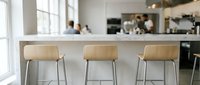 Three wooden bar stools with metal legs positioned at kitchen counter in modern home interior with blurred figures and appliances in background.