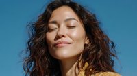 Young woman with curly hair enjoying sunshine against blue sky background, eyes closed with peaceful expression and natural makeup.