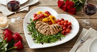 Grilled chicken breast with roasted vegetables and arugula on white plate, romantic dinner setting with red roses and wine glasses on rustic wooden table.