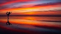 Woman practicing yoga dancer pose on beach at sunset with vibrant orange sky reflecting on wet sand creating peaceful meditation scene for wellness.