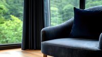 Cozy gray armchair with navy blue velvet cushion near window overlooking lush green forest view, creating serene home interior atmosphere with natural daylight.