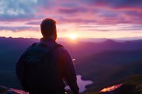 Male hiker watching dramatic purple sunset over mountain lake, silhouetted figure with backpack observing majestic wilderness landscape from summit viewpoint.