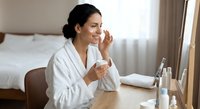 Young Hispanic woman in white bathrobe applying skincare cream at vanity mirror in modern bedroom. Morning beauty routine and self-care concept.