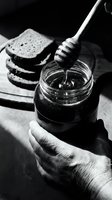 Hand holding glass jar of honey with wooden dipper, sliced bread in background, monochrome food styling for culinary, breakfast and organic product promotion.