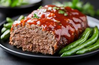 Homemade meatloaf glazed with tomato sauce, served on black plate with fresh green beans, garnished with herbs, close up view of comfort food meal.