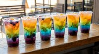 Five colorful rainbow layered drinks with straws arranged on wooden table at outdoor cafe terrace during golden hour sunset lighting.